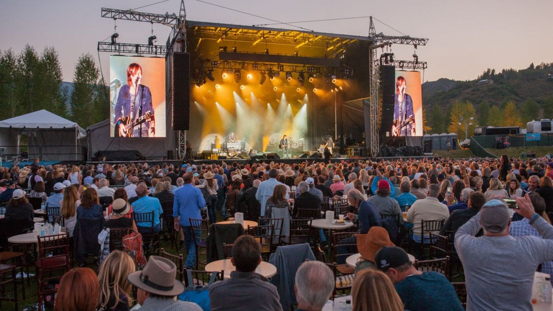 large audience watching a concert with light show and large video screens on either side of a stage with mountains and trees beyond 