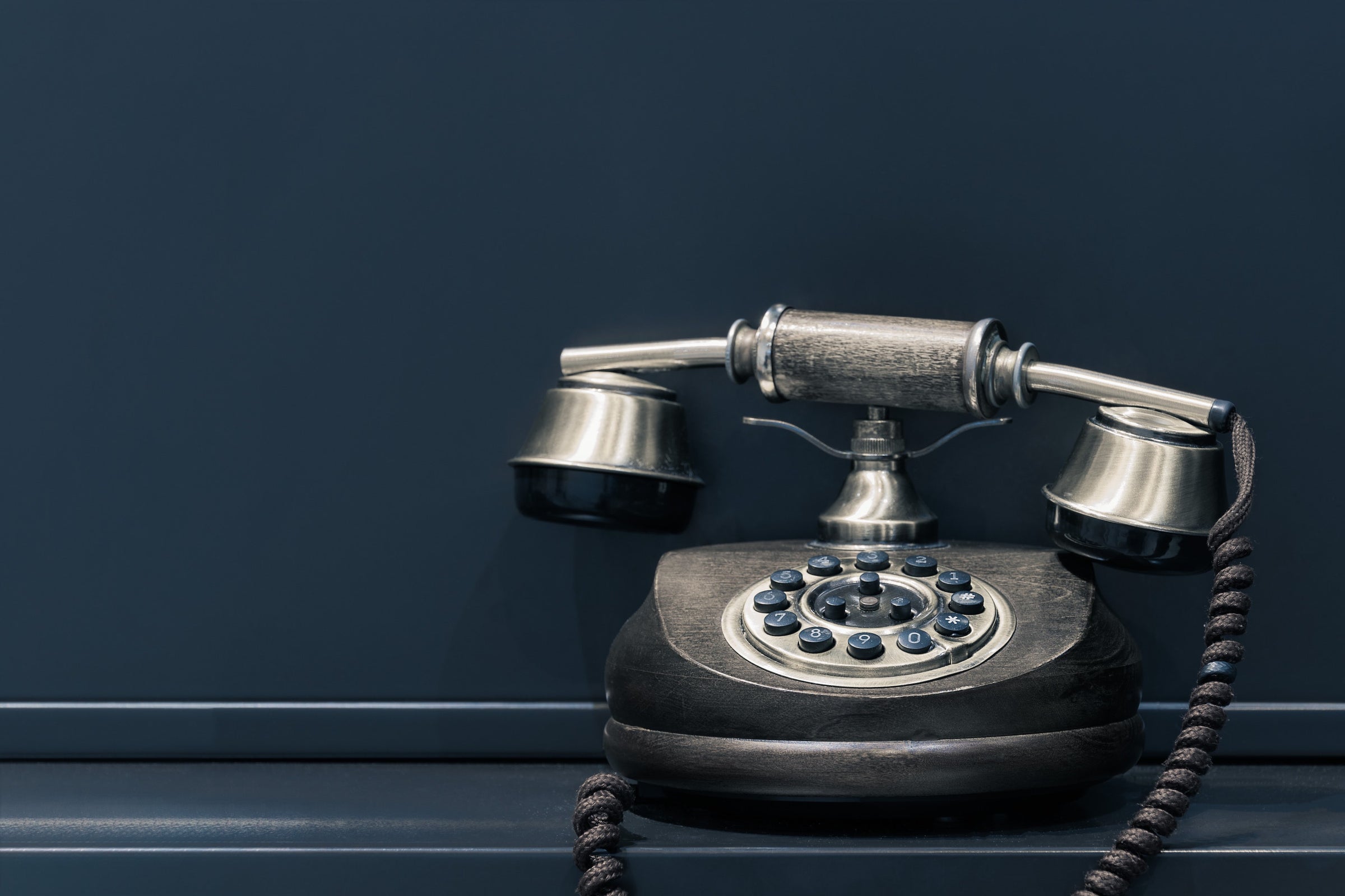 stainless steel finish rotary style telephone with blue buttons, gray cord in front of a slate blue background on a slate blue surface