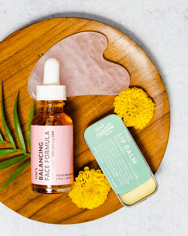 wooden platter with rose quartz guasha, yellow flowers, glass bottle of face oil with pink label, tin of lip balm with mint green label