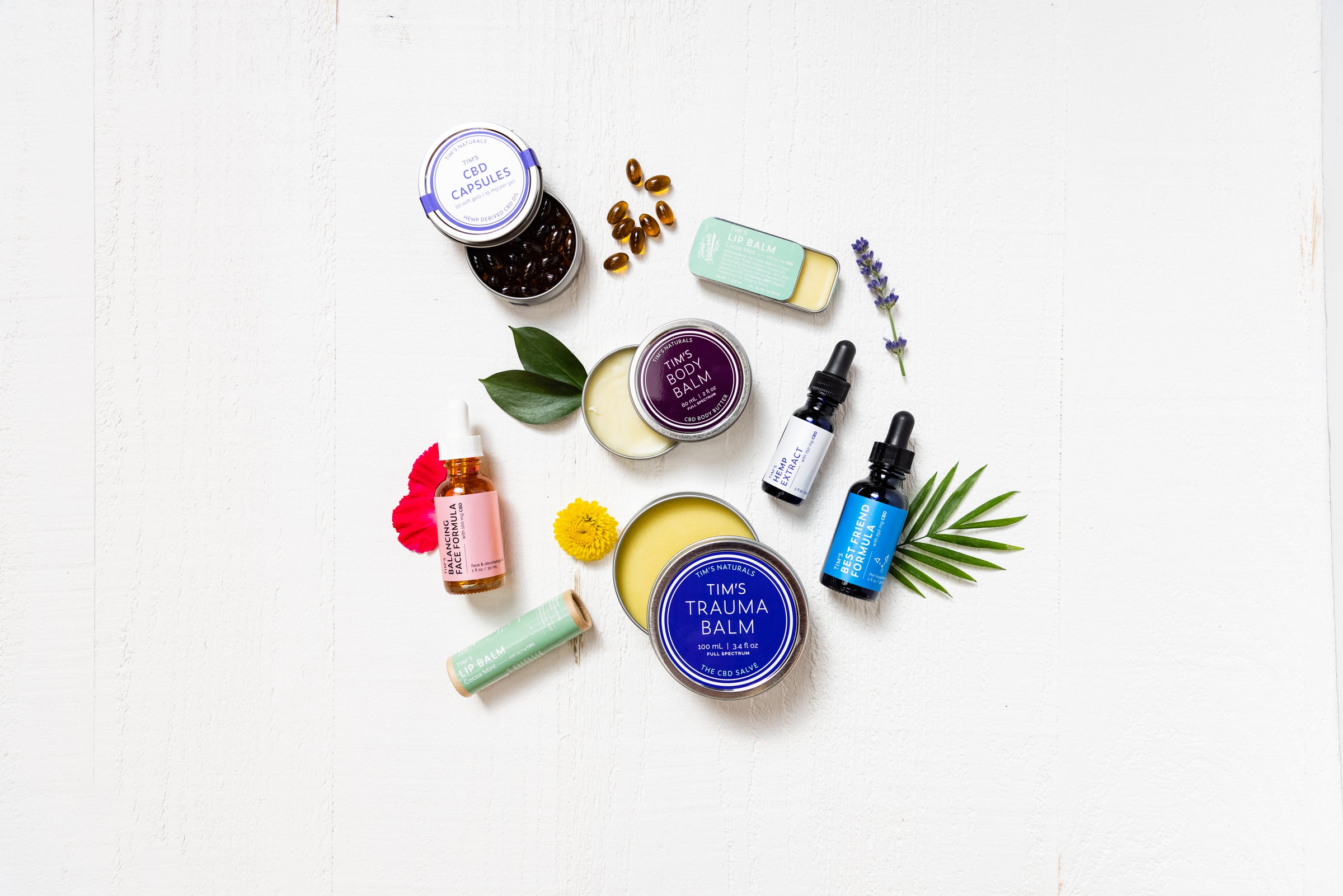Tim's Naturals products with natural, fair-trade, non-GMO, organic ingredients alongside them. Tim's Best Friend Formula, Tim's Lip Balm, Tim's CBD Capsules, Tim's Trauma Balm, Tim's Body Balm, Tim's Balancing Face Formula 